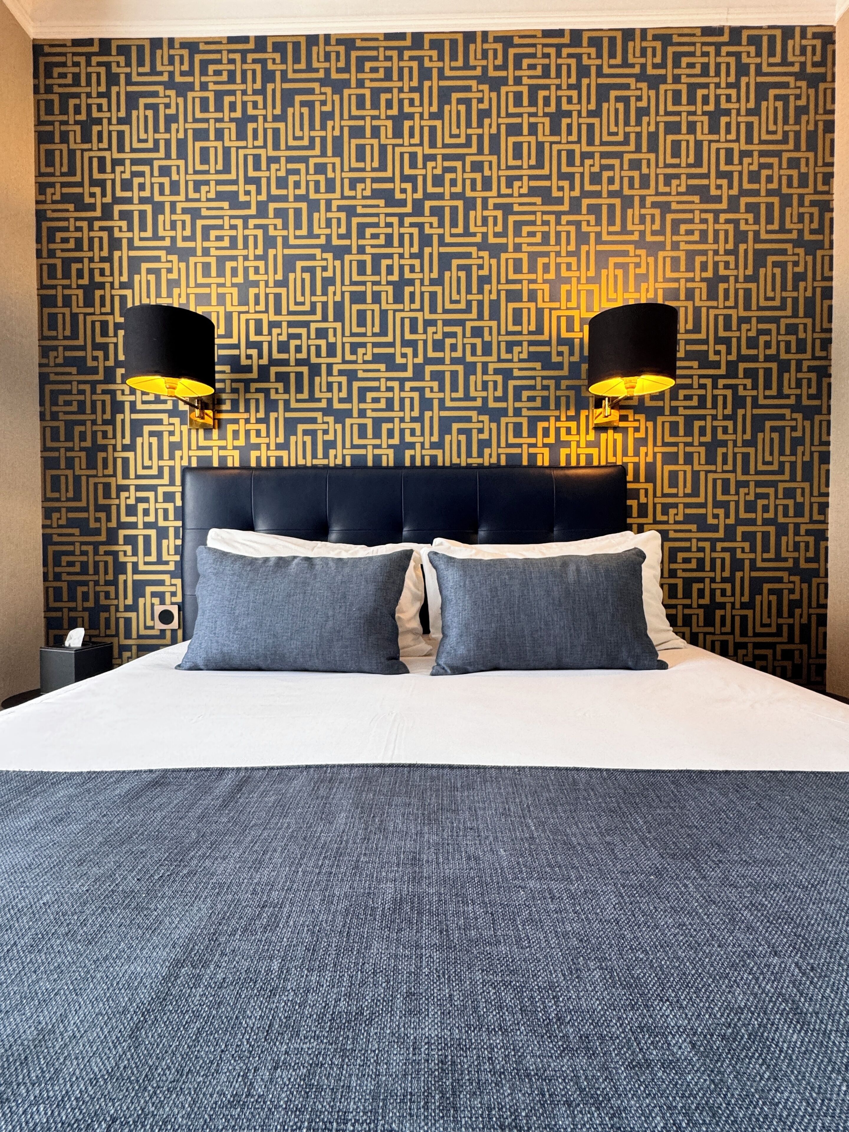 Habitación de hotel con papel tapiz estampado, cabecero acolchado, dos lámparas de noche y una cama bien hecha con ropa de cama gris.
