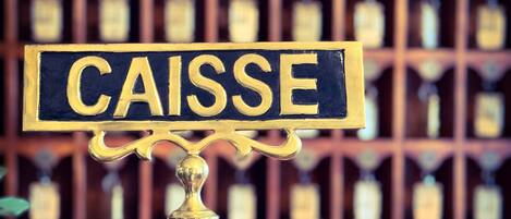 A brass sign with the word "CAISSE" on a metal pole, with a blurred background of wooden cabinets and bottles.