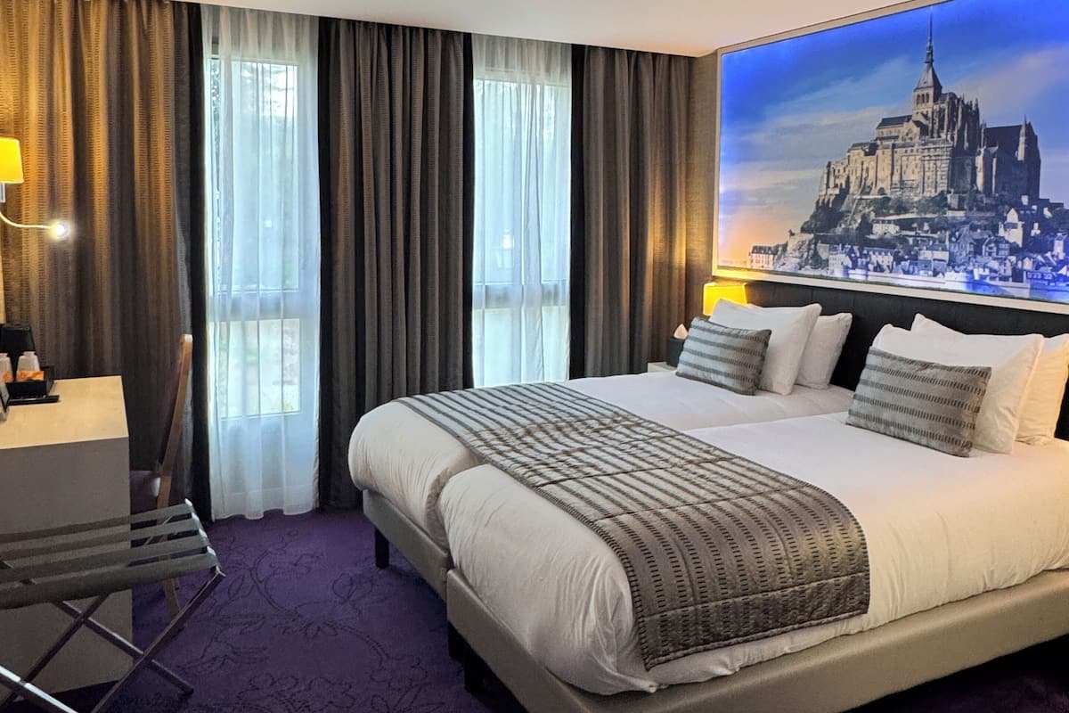 A hotel room with a large bed, a desk, and a chair. There is a window with curtains and a painting of a cityscape on the wall.