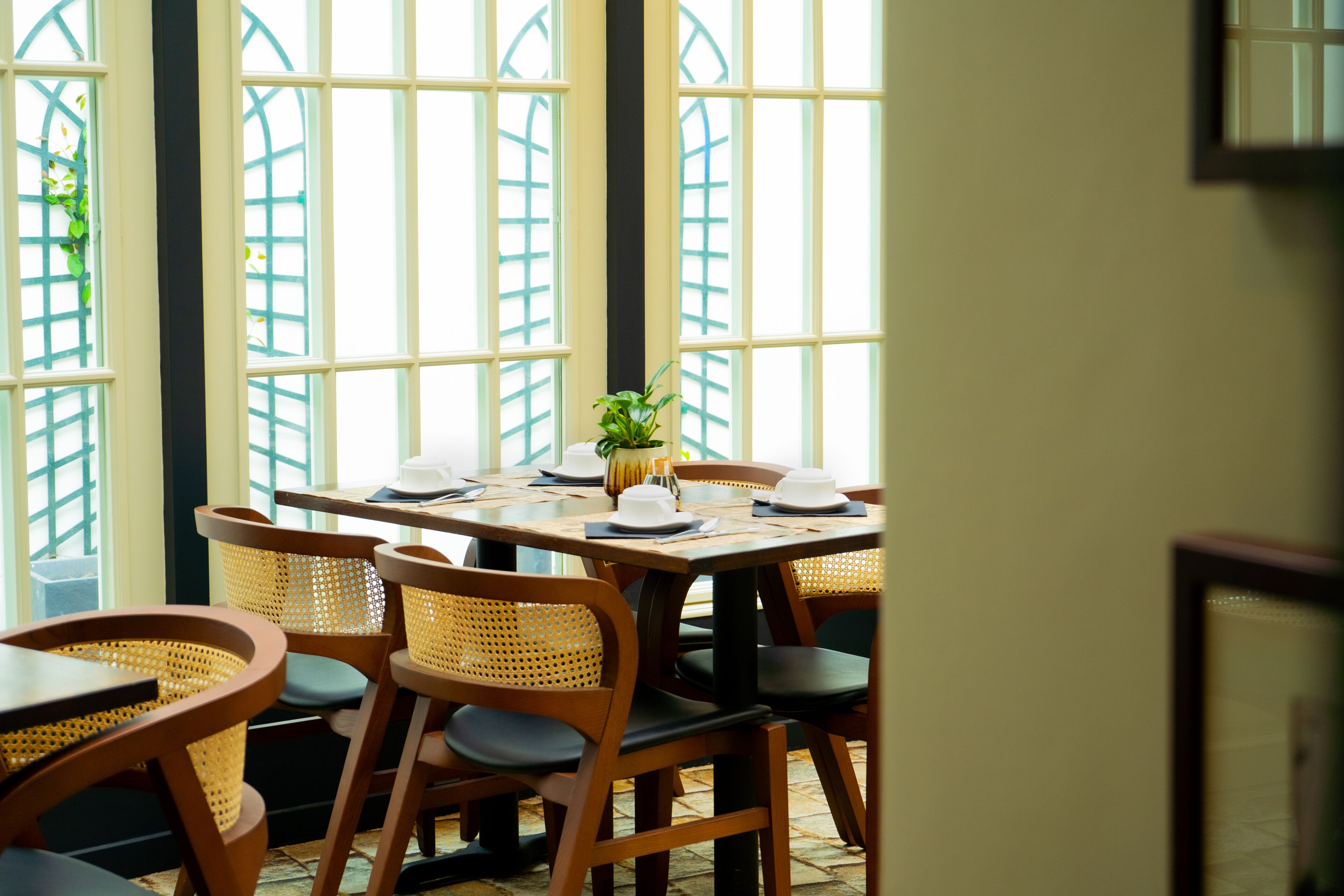 Uma área de refeições com uma mesa posta para quatro pessoas, rodeada por cadeiras de vime, e grandes janelas com vista para o verde lá fora.