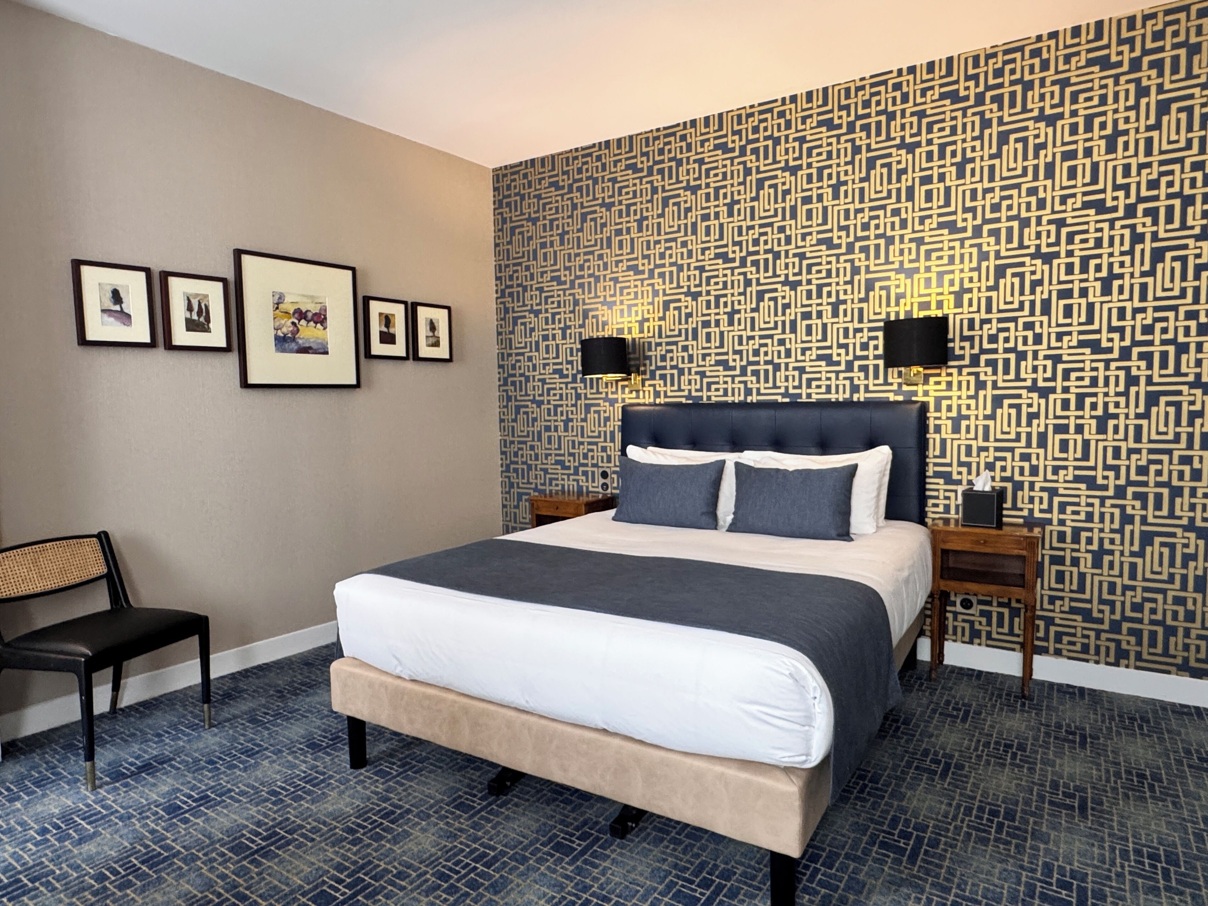 Quarto de hotel com uma cama grande, uma cadeira, uma mesinha de cabeceira e uma parede com padrão geométrico.