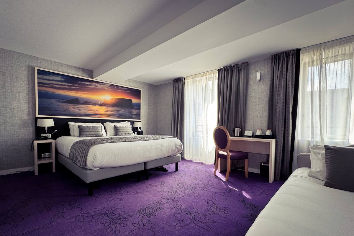 A hotel room with a large bed, a desk, and a chair. There is a painting on the wall and a window with curtains.