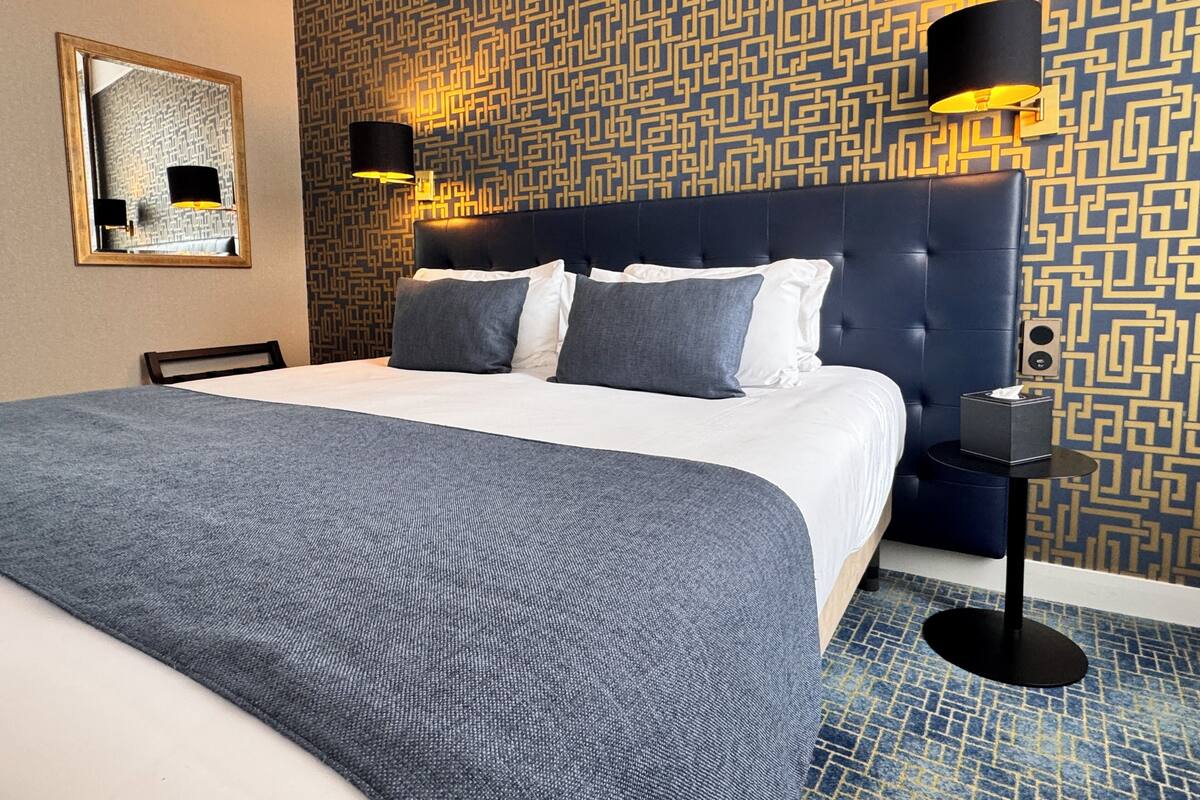 A hotel room with a large bed, a headboard, two wall-mounted lamps, a small table with a tissue box, and a patterned wall.