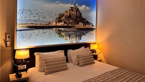 A hotel room with a large bed, two bedside lamps, a nightstand, and a wall-mounted TV displaying a scenic view of a castle and river.