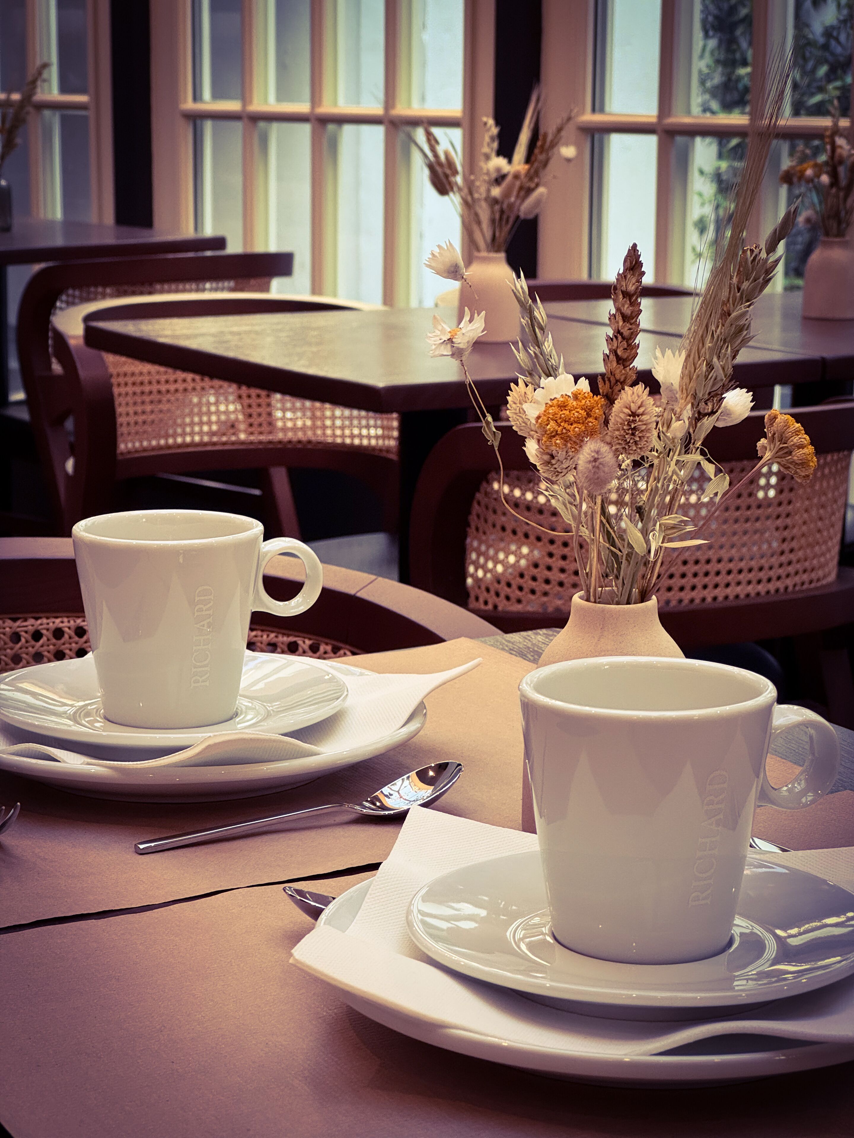 A table set with white cups and saucers, a vase with dried flowers, and a fork and spoon on a napkin.