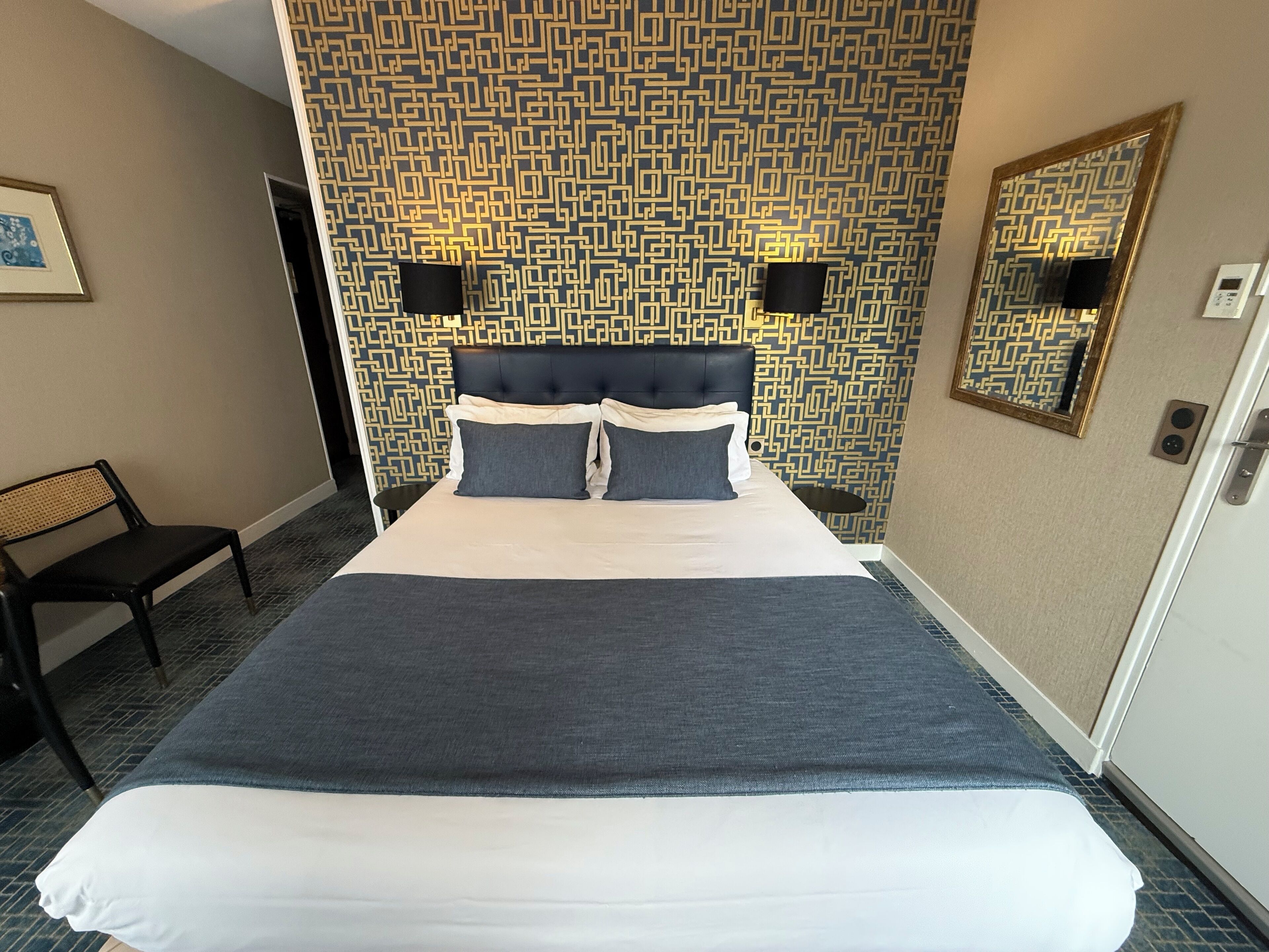 Une chambre d’hôtel avec un grand lit, une tête de lit, deux lampes fixées au mur, une table de chevet, une chaise et un miroir sur le mur.