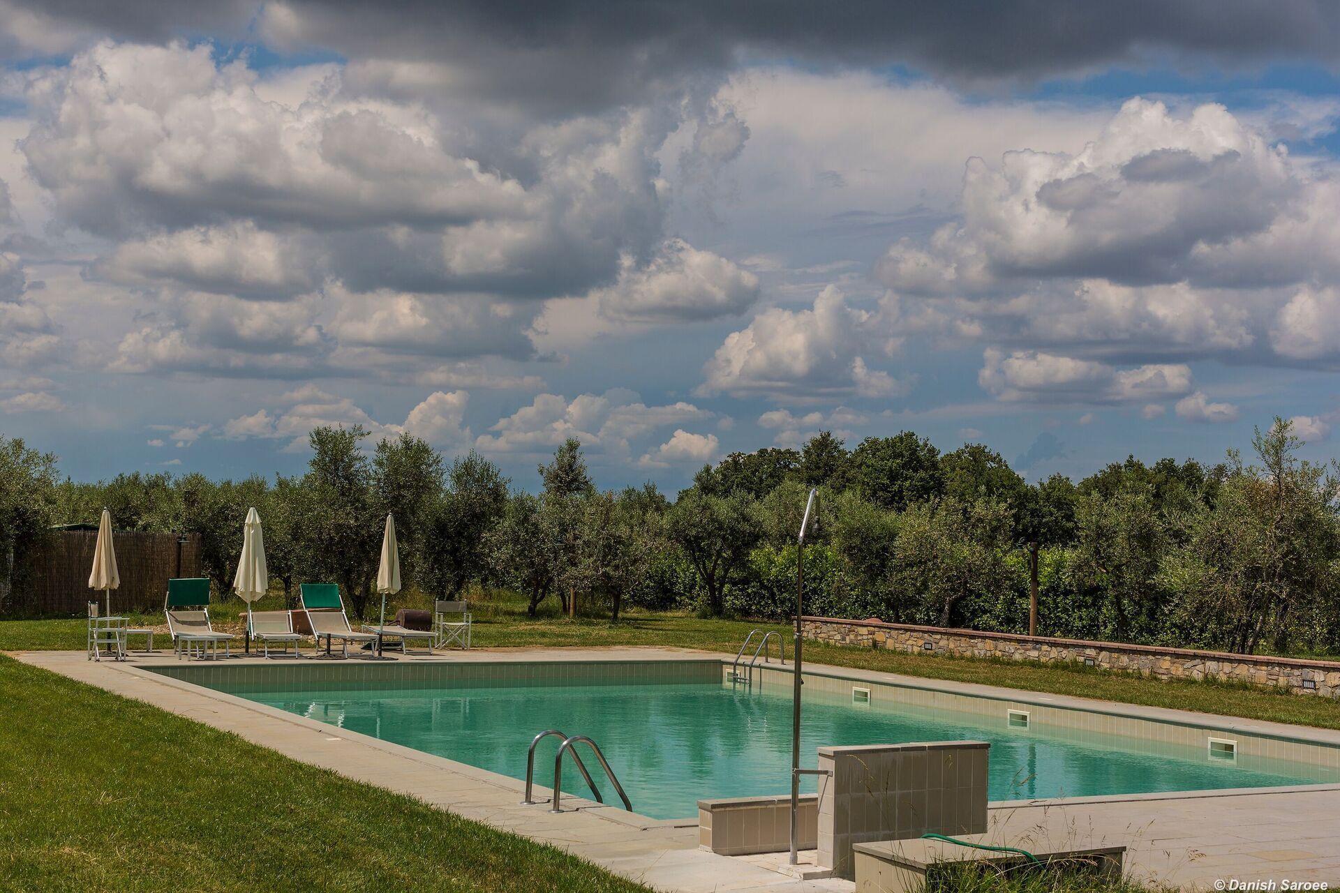 Une piscine rectangulaire bordée de pierres, entourée de pelouse et d’arbres.
