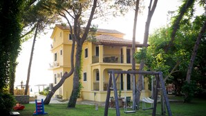 A yellow building with a balcony, surrounded by trees and a swing set. - Zarkadis Beach Apartments (Zakynthos)