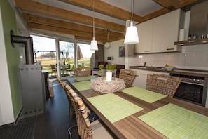 A modern kitchen with a long dining table, wicker chairs, and pendant lights.
