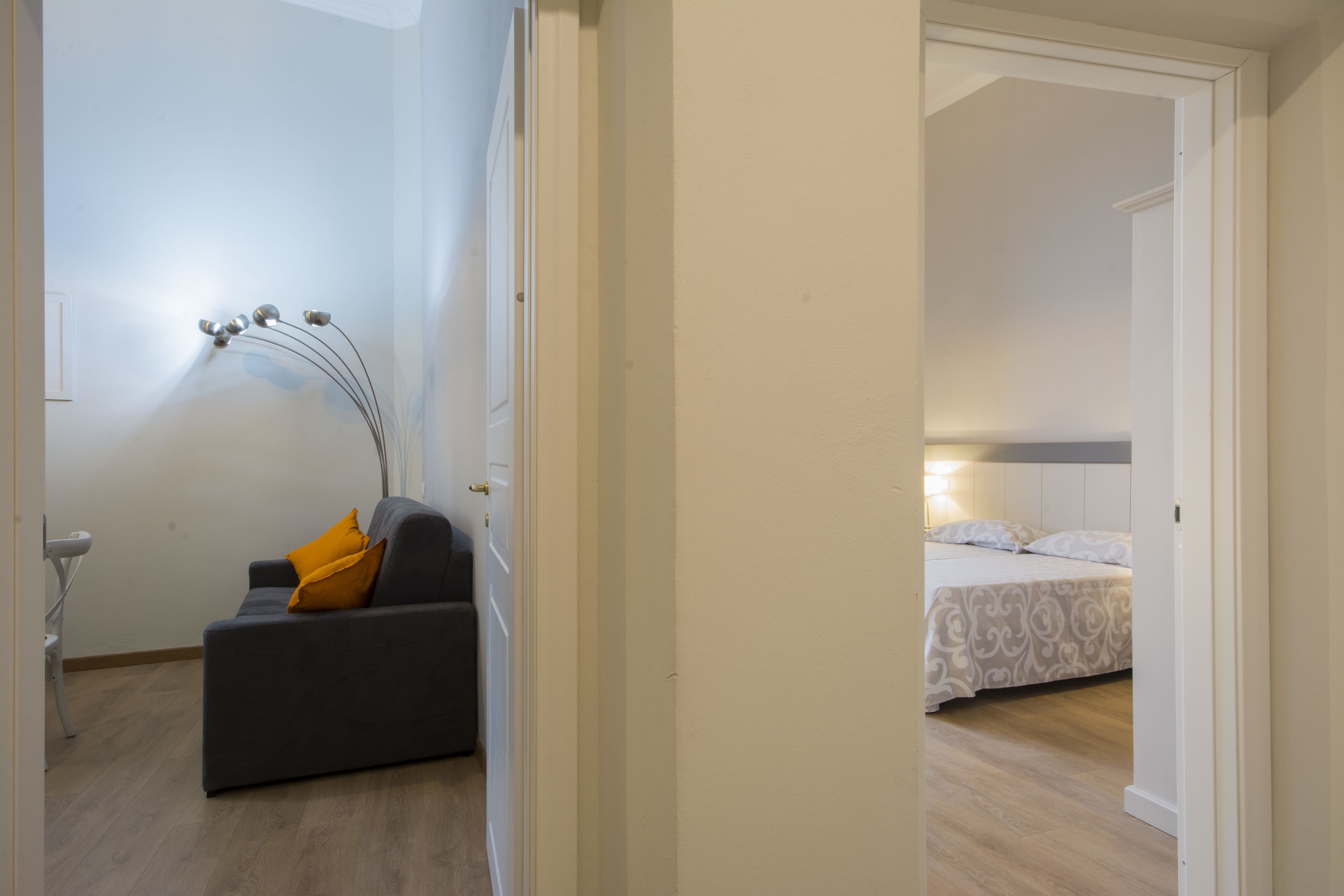 A room with a grey sofa, a yellow pillow, a bedside table, and a bed with patterned bedding.