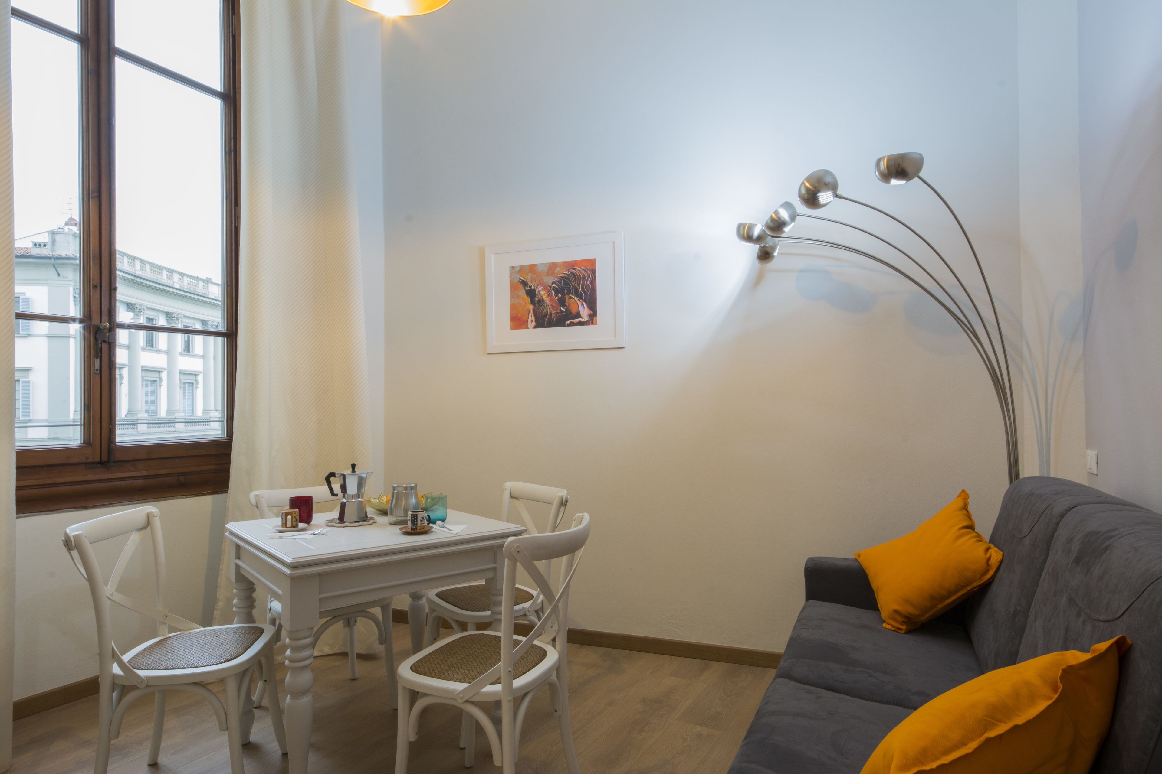 A living room with a dining table, chairs, a grey sofa with yellow cushions, a framed picture on the wall, and a modern floor lamp.