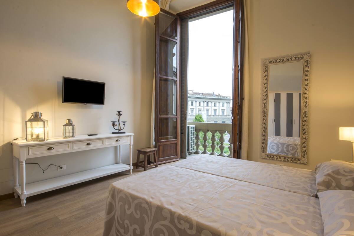 Une chambre à coucher comprenant un lit, une commode, une télévision, un balcon avec vue sur les immeubles et un miroir.