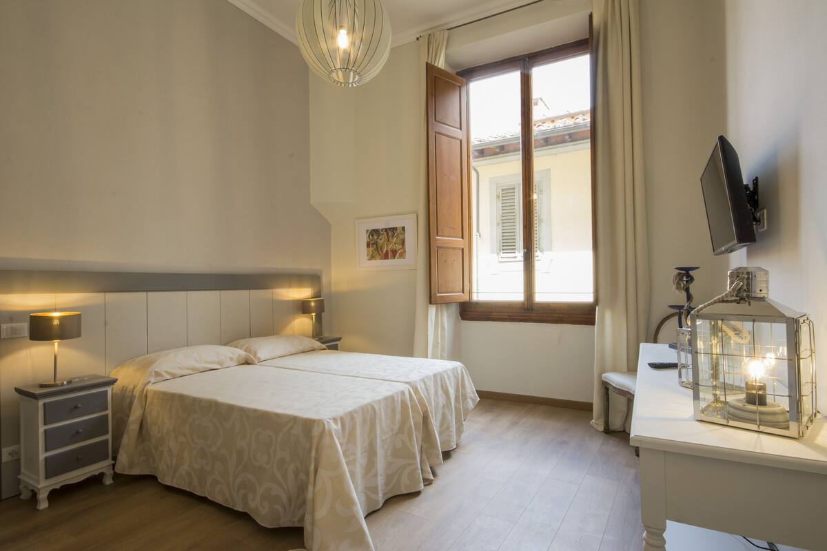 Une chambre avec deux lits, une table de chevet, un bureau avec une lampe, une fenêtre avec des rideaux et un téléviseur fixé au mur.