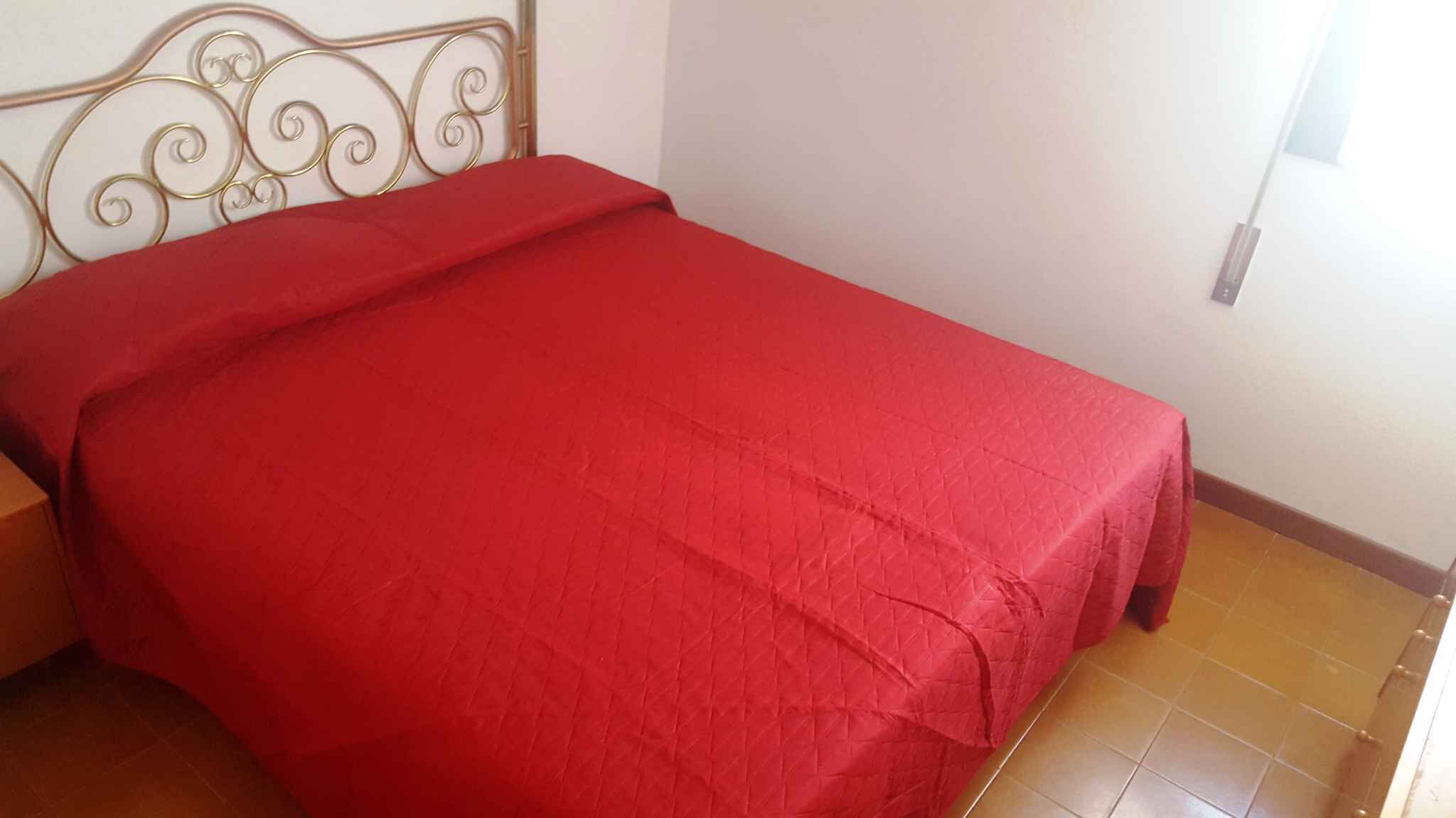 Un lit simple avec une couvre-lit rouge, dans une pièce dont le sol est carrelé et qui est dotée d’une tête de lit en métal.