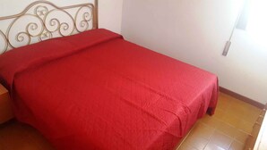 Een enkel bed met een rode sprei in een kamer met een betegelde vloer en een metalen hoofdbord.