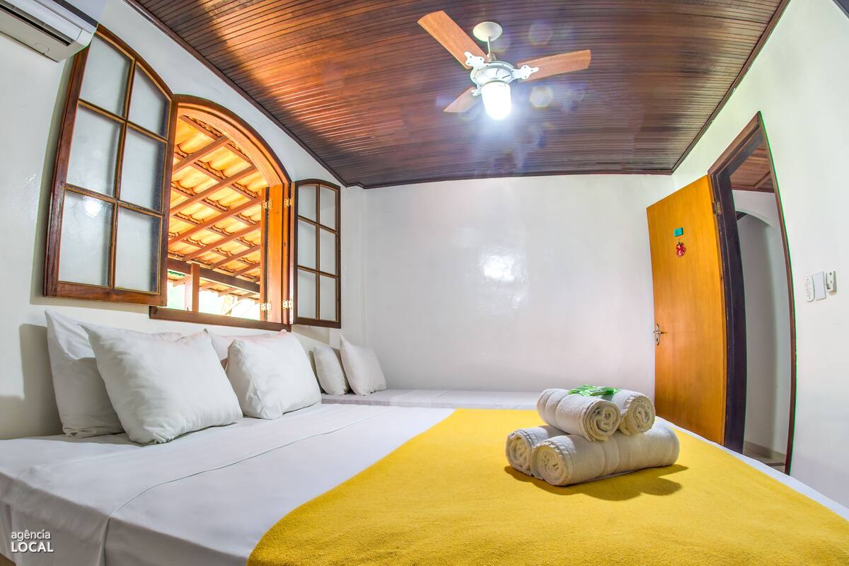 A hotel room with a bed, a ceiling fan, a wooden door, and a window with a wooden frame.