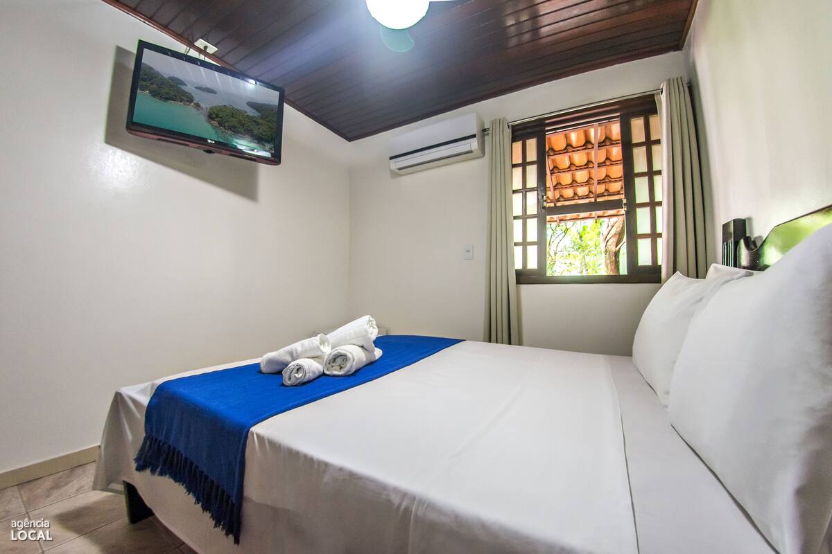 A hotel room with a bed, a TV mounted on the wall, a window with curtains, and a view of greenery outside.