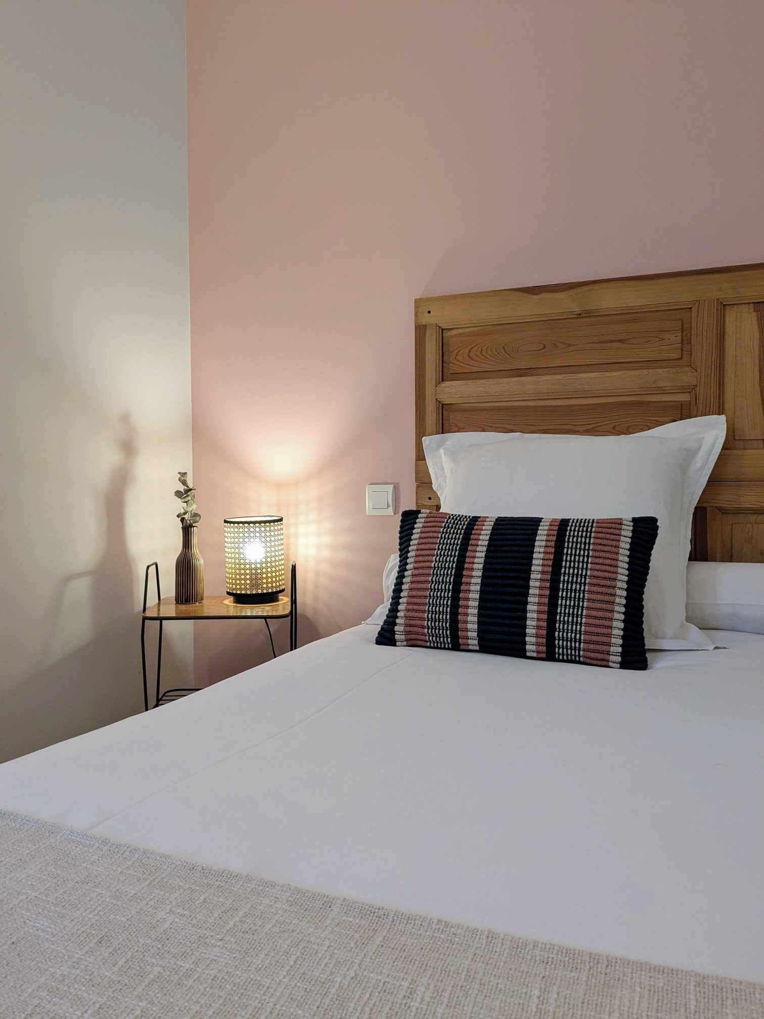 A neatly made bed with a striped pillow, a bedside table with a lamp, and a wall-mounted switch.