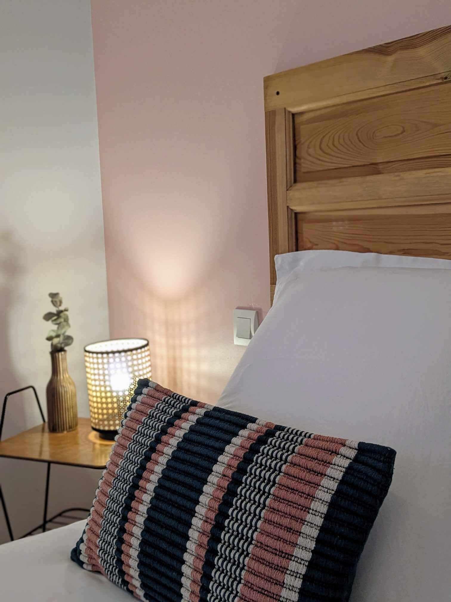 Una cama bien hecha con cabecera de madera, una lámpara de noche y un cojín estampado.