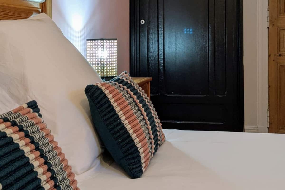 Un dormitorio con una cama, almohadas, un cabecero de madera, un armario negro y una mesita de noche con una lámpara.