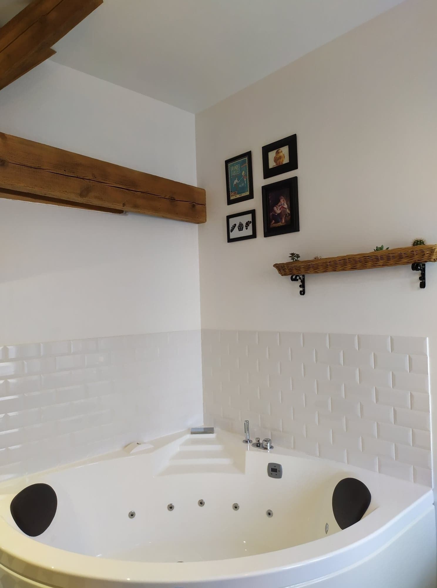 A bathroom with a large bathtub, wooden beams, a white tiled wall, and a shelf with decorative items.