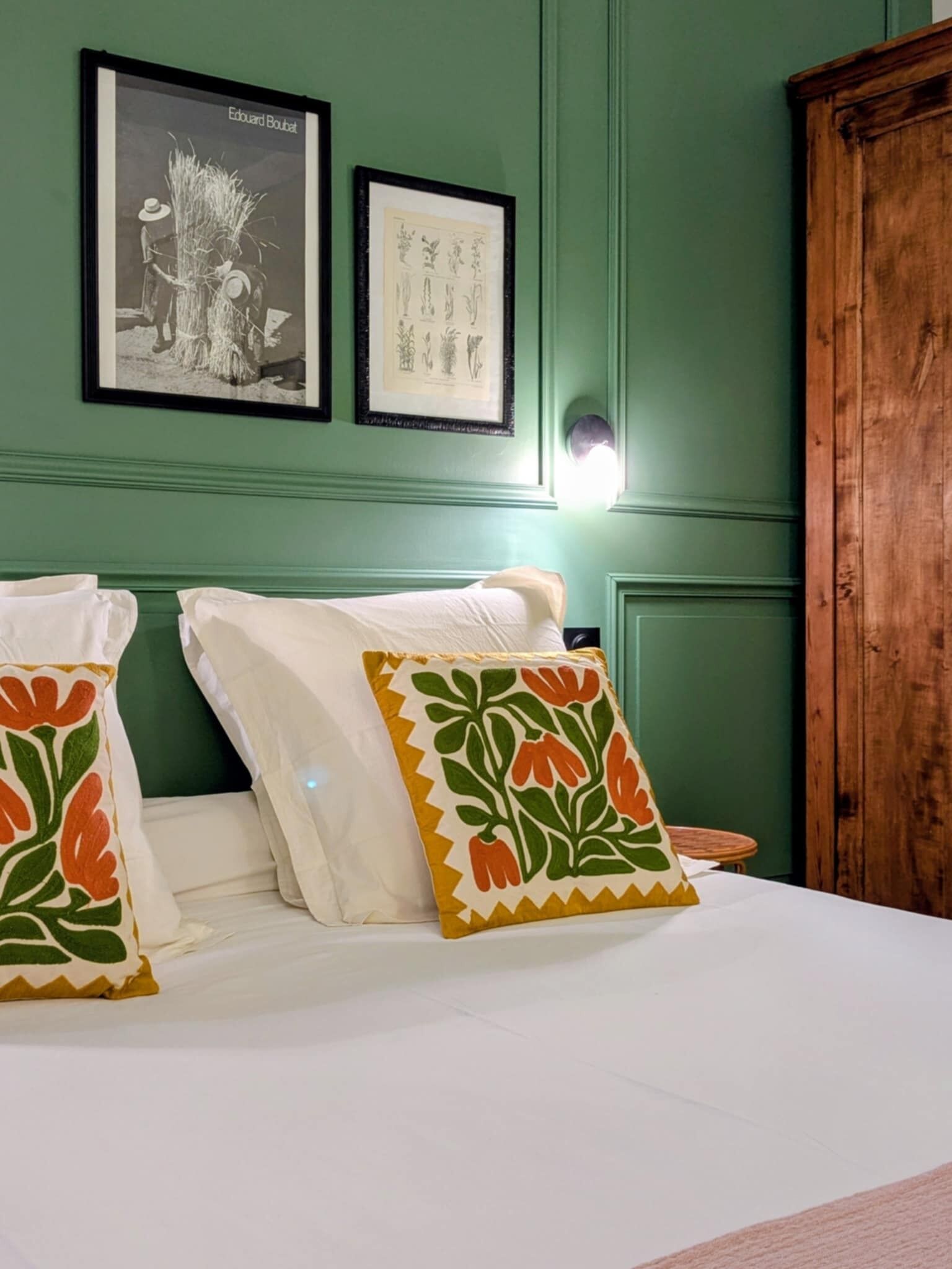 Una habitación con una cama, dos almohadas, un armario de madera e ilustraciones botánicas enmarcadas en la pared.