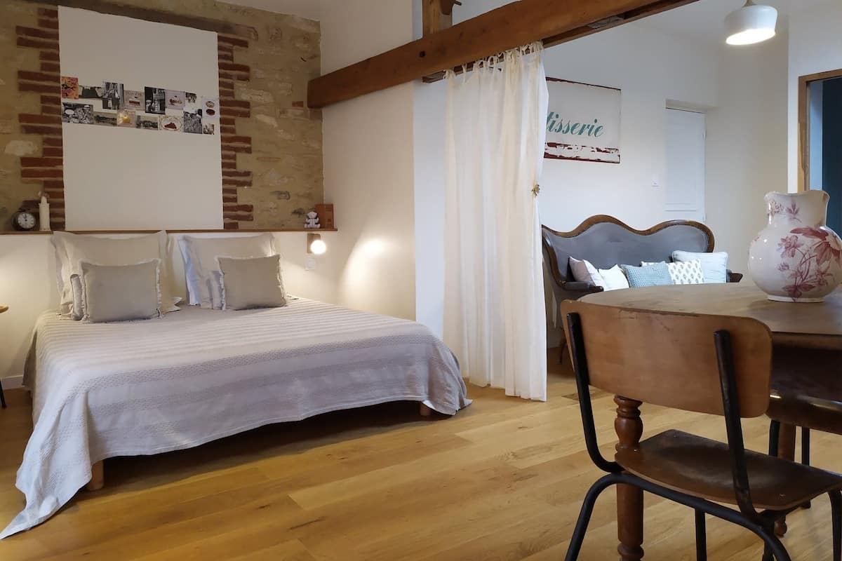 Un dormitorio con cama, suelo de madera, una mesa de comedor con sillas, pared de ladrillo y techo con vigas a la vista.