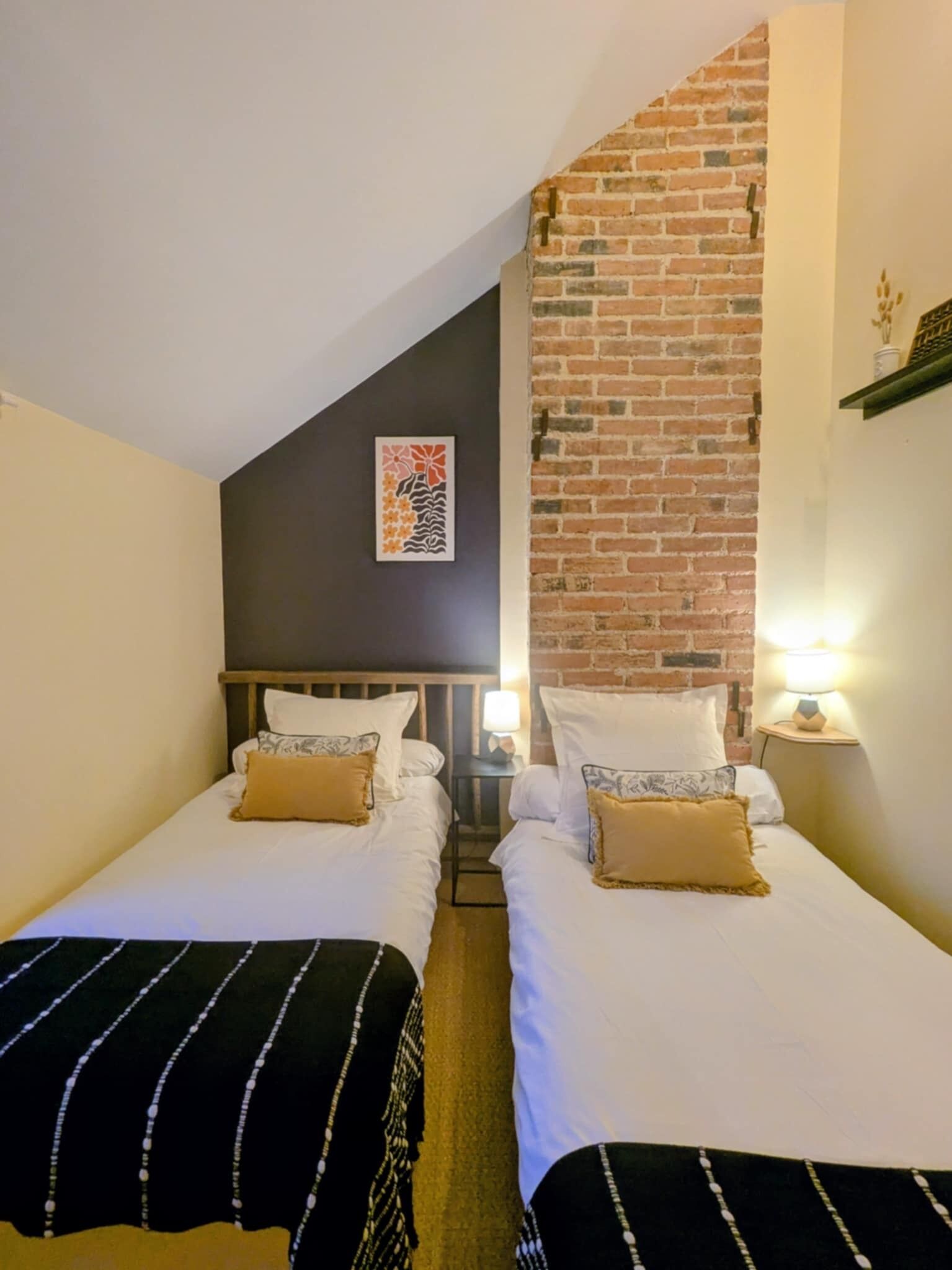 A room with two single beds, a brick wall, a framed artwork, and a small table with a lamp.