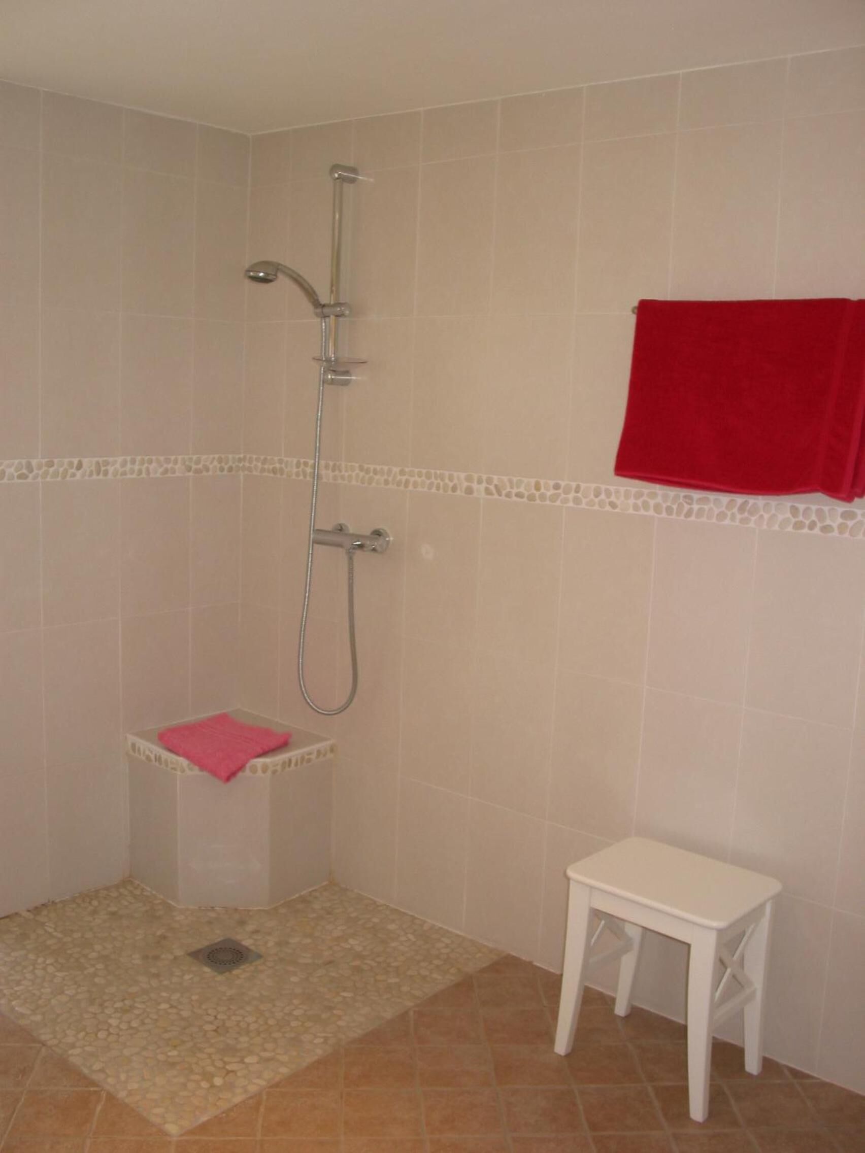 A shower with a handheld showerhead, a small shelf with a pink towel, and a white stool.