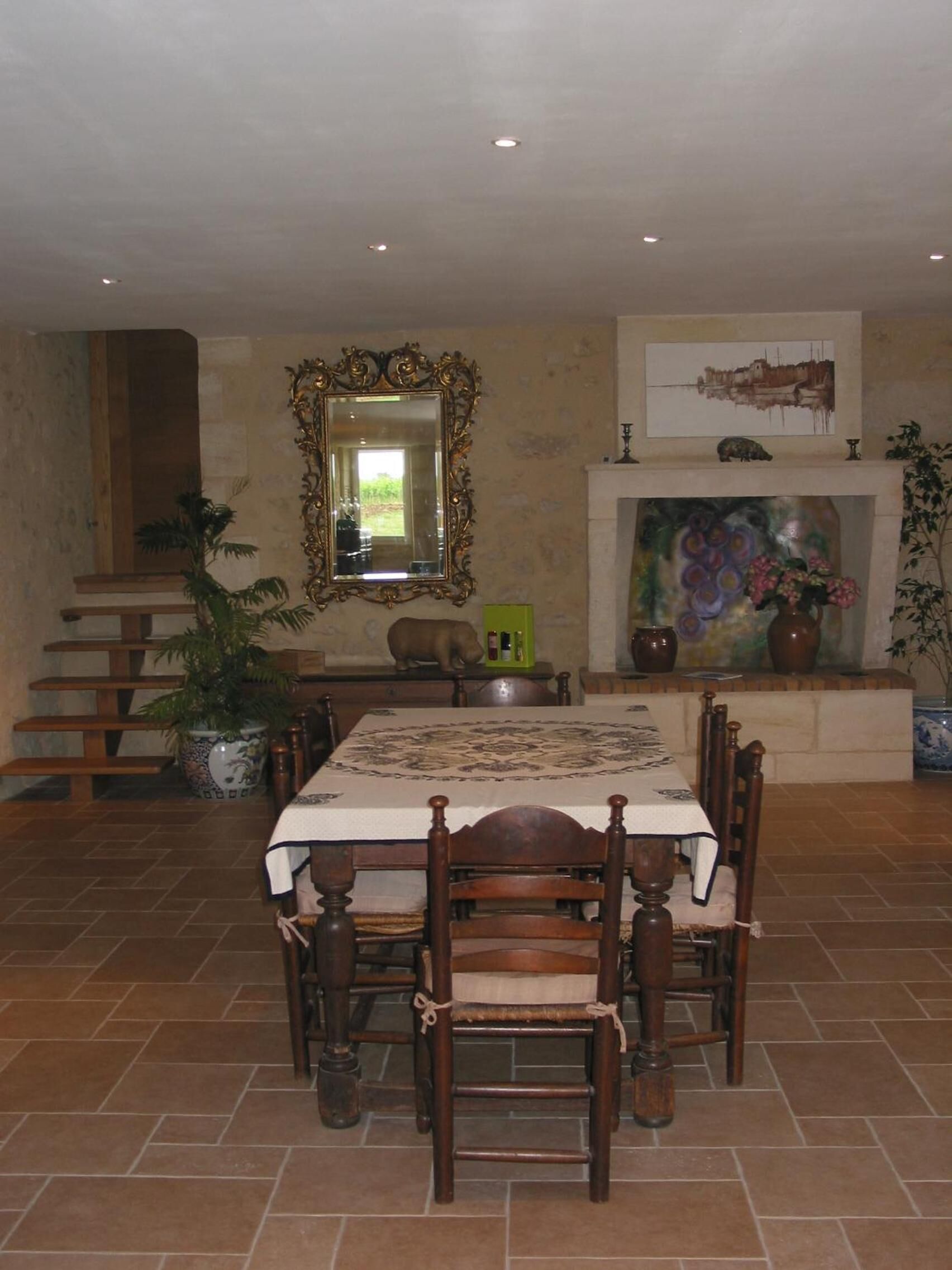 A dining area with a table set for six, surrounded by wooden chairs. A large mirror hangs above the table, and a fireplace with a colorful mural is visible in the background.
