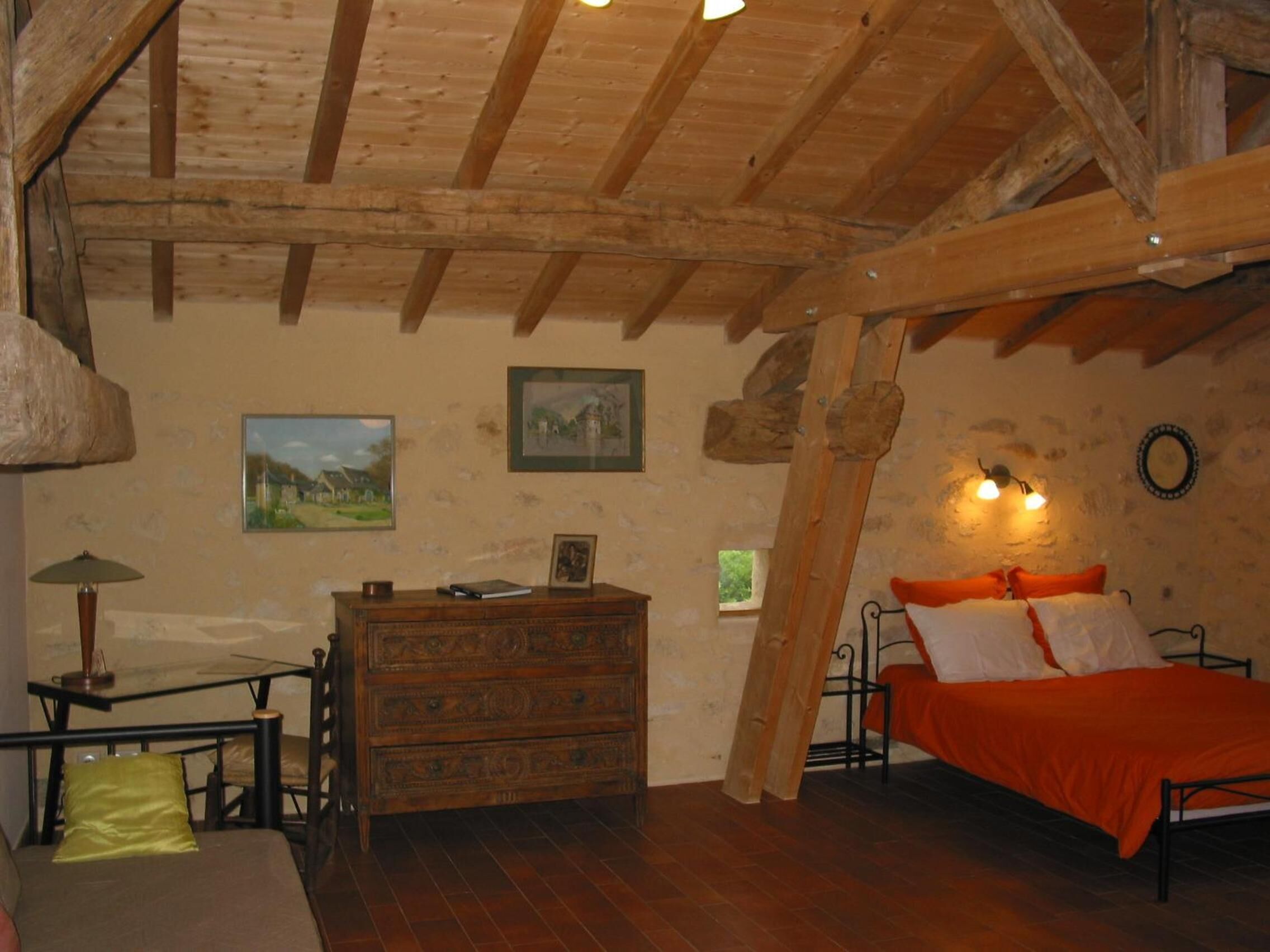 Ein rustikales Schlafzimmer mit freiliegenden Holzbalken, einem hölzernen Schminktisch und einem Bett mit orangefarbener Bettwäsche, sowie zwei Wandleuchten.