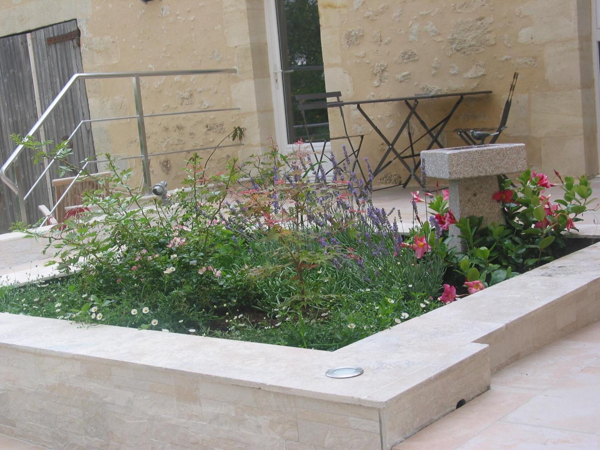 Een tuin met diverse planten en bloemen, een metalen hekwerk en een stenen bank.