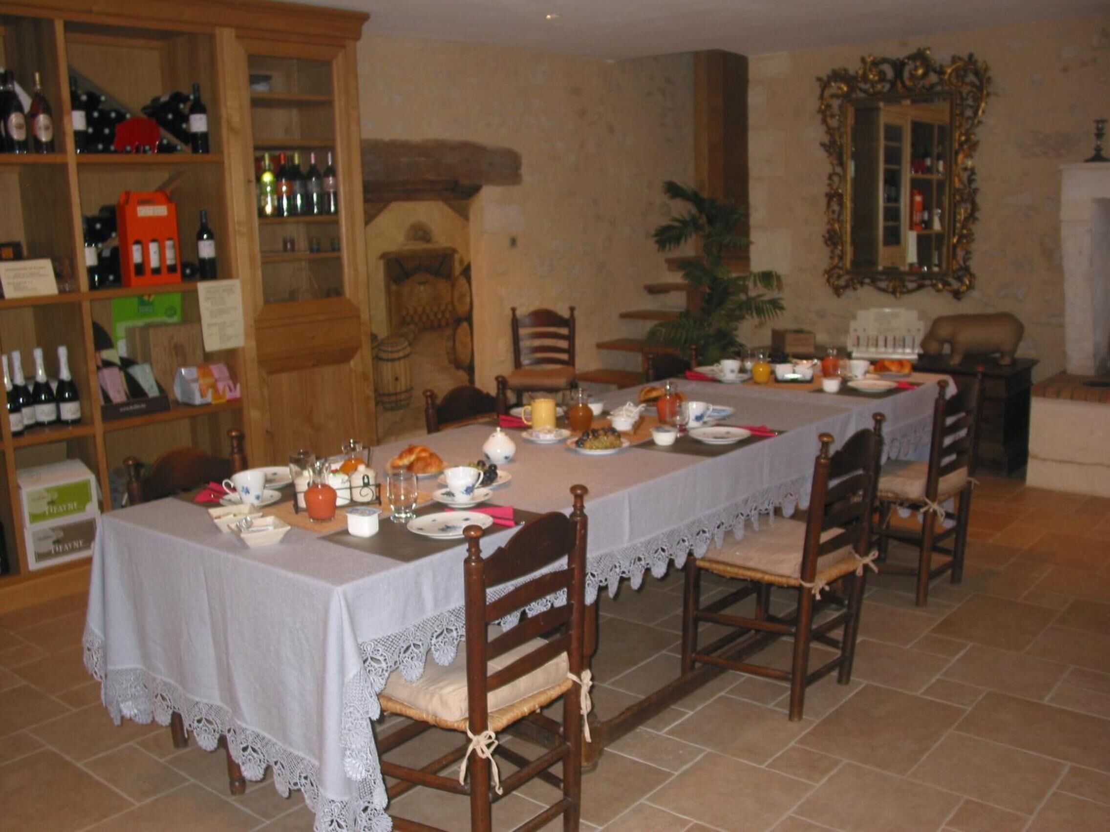 A dining room with a long table set for a meal, surrounded by wooden chairs. A bookshelf with wine bottles is visible to the left.