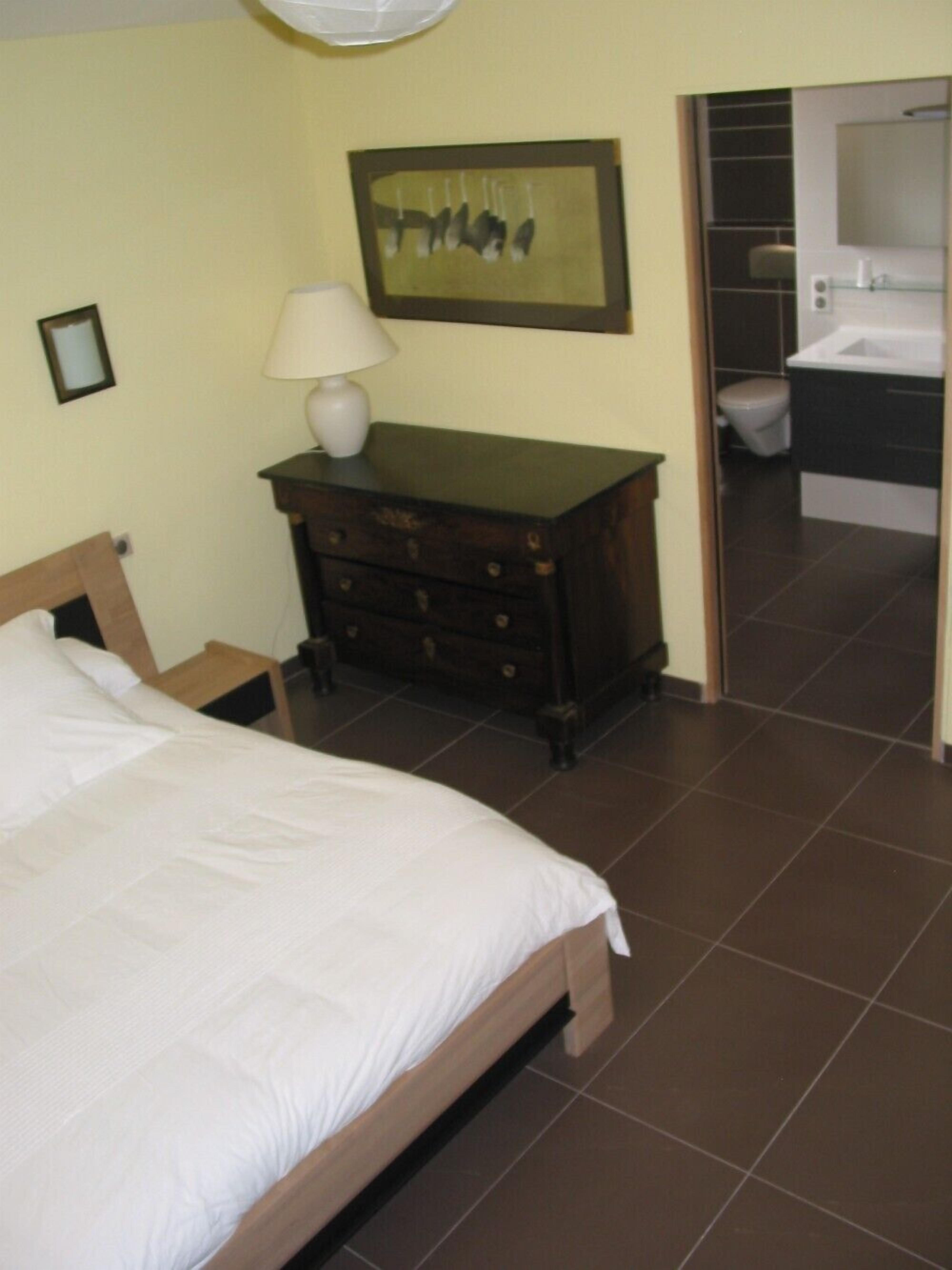 A bedroom with a bed, a dresser, a nightstand, a framed picture, and an open door to a bathroom.