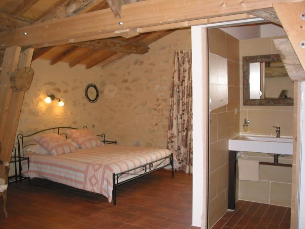 A bedroom with a bed, a wooden ceiling, a bathroom with a sink and mirror, and a stone wall.