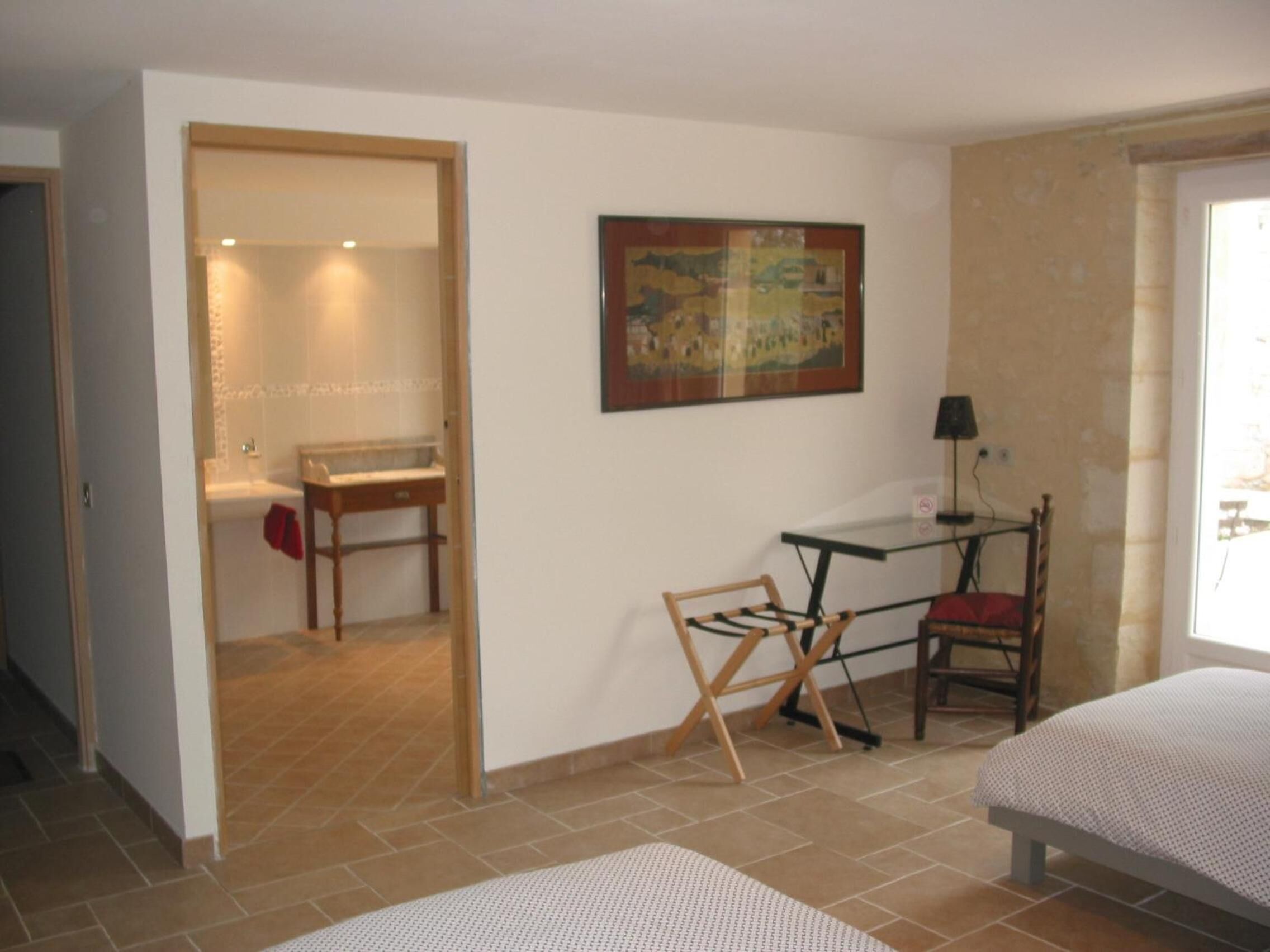 A room with a bed, a desk, a chair, a painting, and a bathroom visible through an open door.