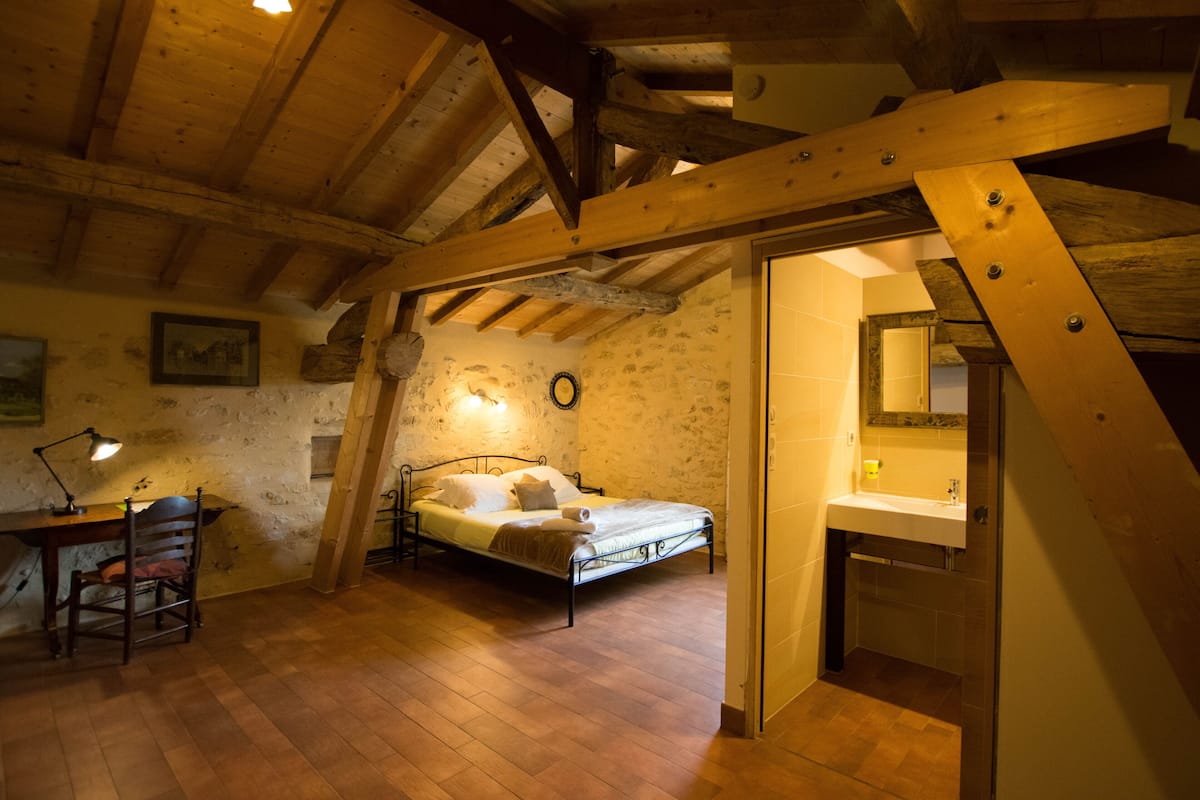 Un dormitorio con techo de madera, una cama, un escritorio con silla, y un baño con lavabo y espejo.