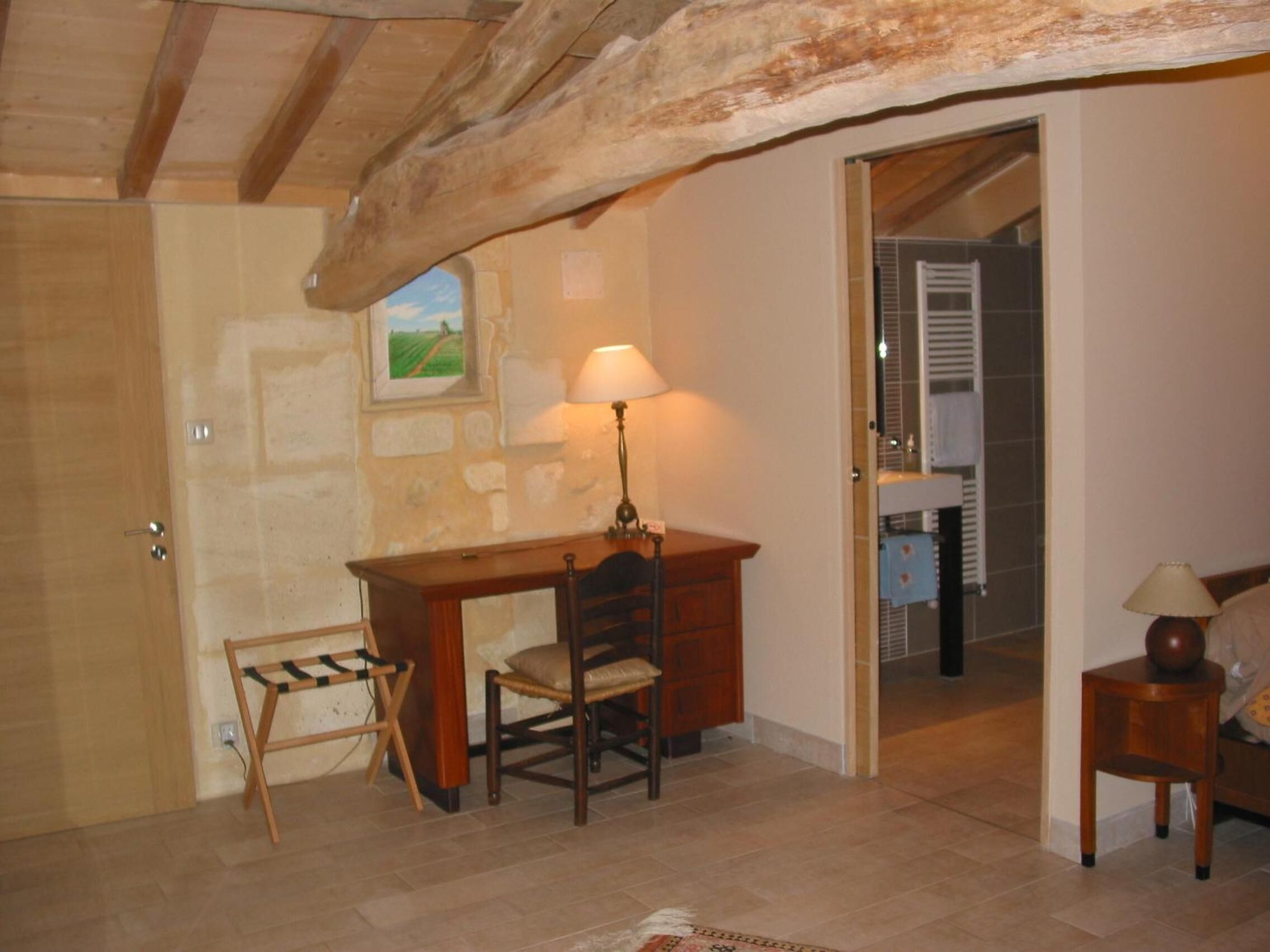 A room with a wooden desk, a chair, a lamp, and a small table. There is a door leading to another room with a tiled floor and a window with blinds.