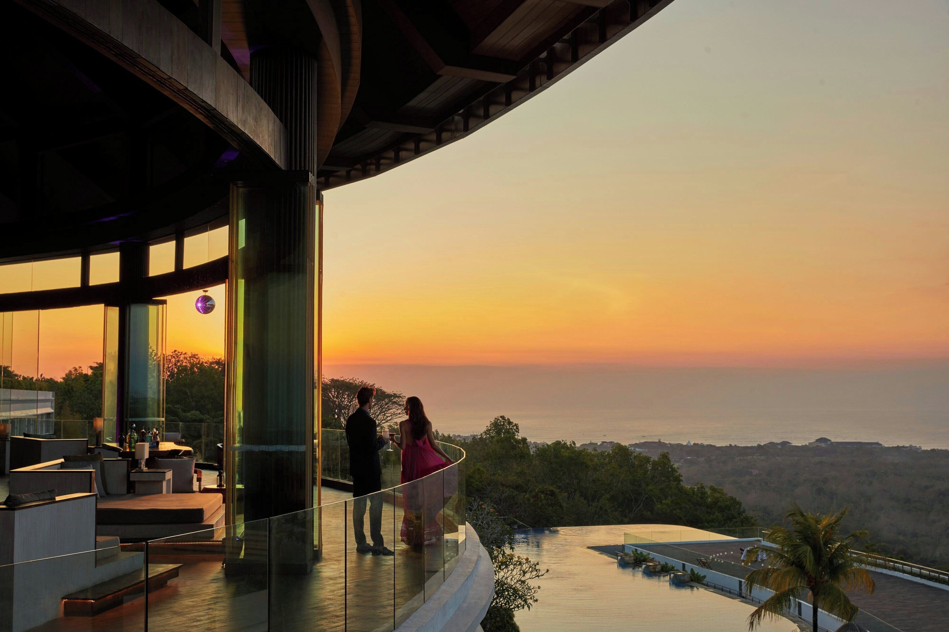 A couple standing on a balcony overlooking a sunset, with a pool and outdoor seating area visible.