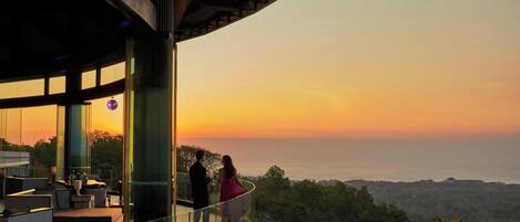 A couple standing on a balcony overlooking a sunset, with a pool and outdoor seating area visible.