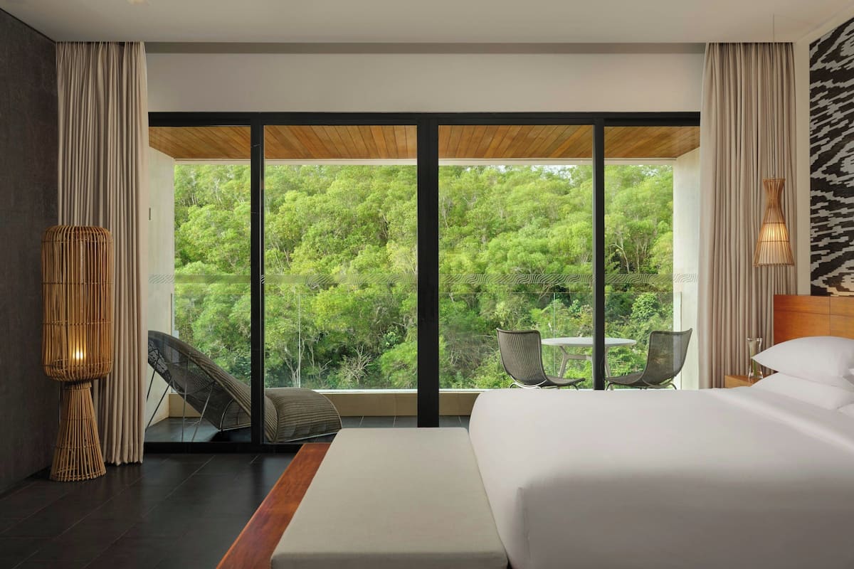 A modern hotel room with a large bed, a seating area with chairs, and a view of lush greenery through floor-to-ceiling windows.