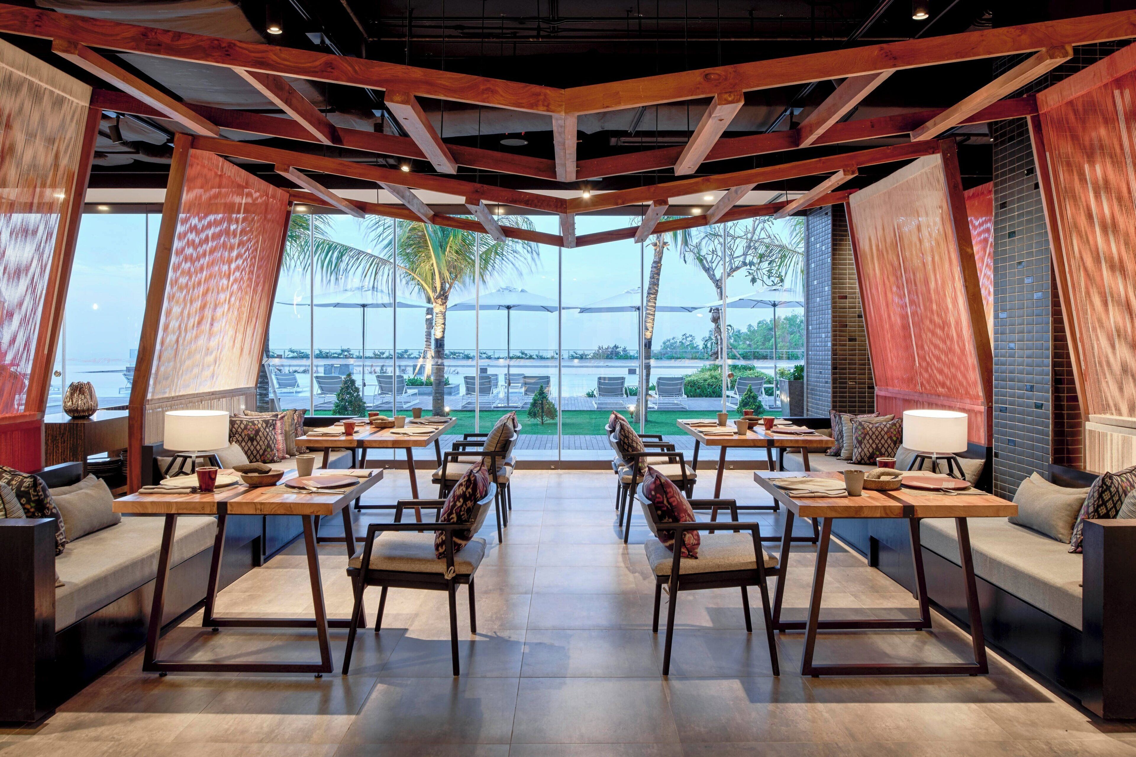 A spacious dining area with high ceilings, wooden beams, and large windows offering a view of palm trees and a body of water. Tables and chairs are arranged for dining.