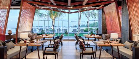 A spacious dining area with high ceilings, wooden beams, and large windows offering a view of palm trees and a body of water. Tables and chairs are arranged for dining.