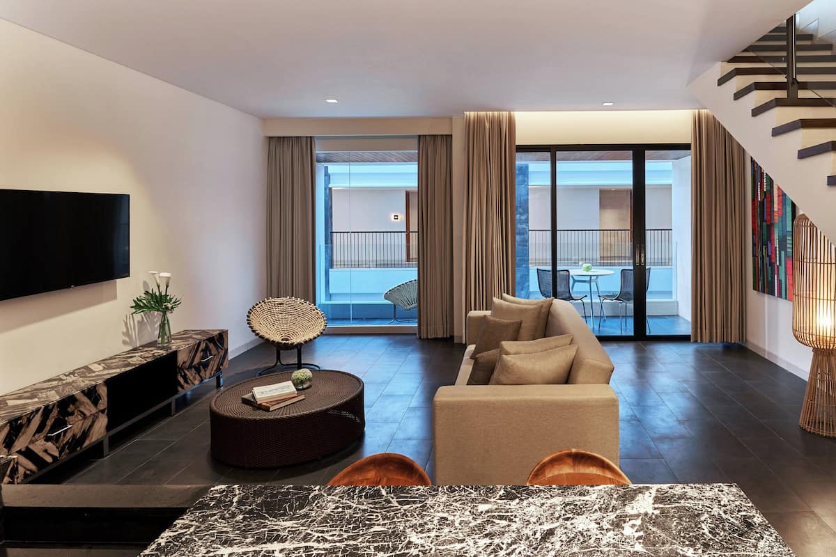 A modern living room with a sofa, coffee table, and TV. There is a balcony with outdoor furniture visible through the glass doors.