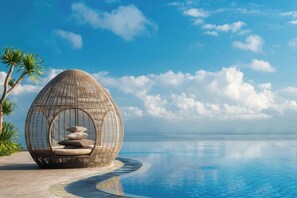 Una silla esférica de mimbre con cojines junto a una piscina, palmeras y un cielo azul despejado.