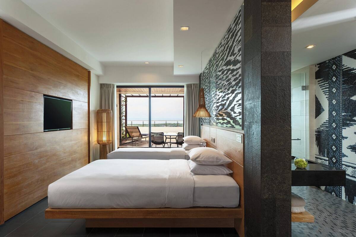 A modern hotel room with a large bed, a TV, and a view of the beach through the window.