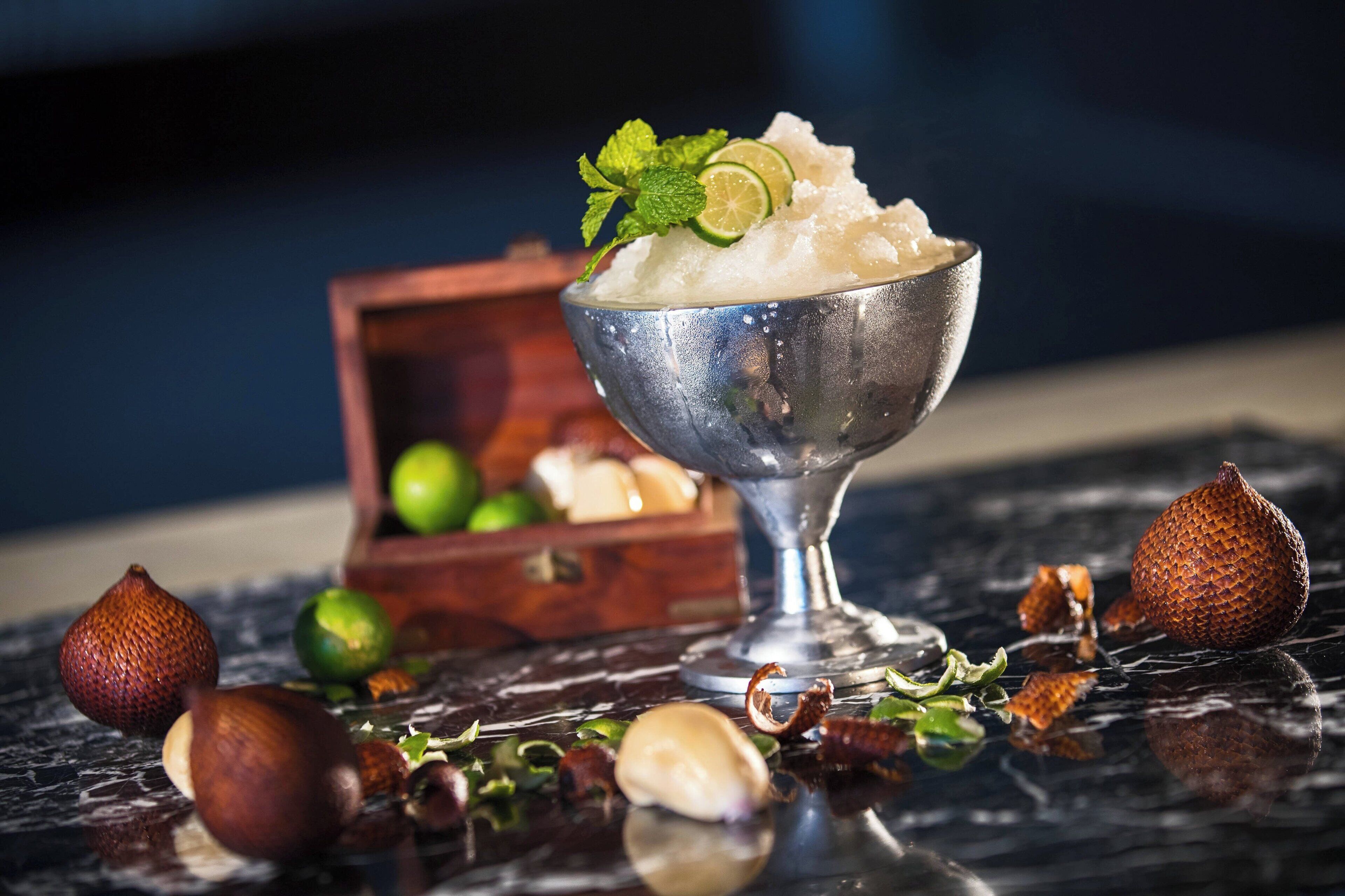 A metal bowl filled with shaved ice, garnished with lime and mint, on a reflective surface with hazelnuts and a wooden box in the background.