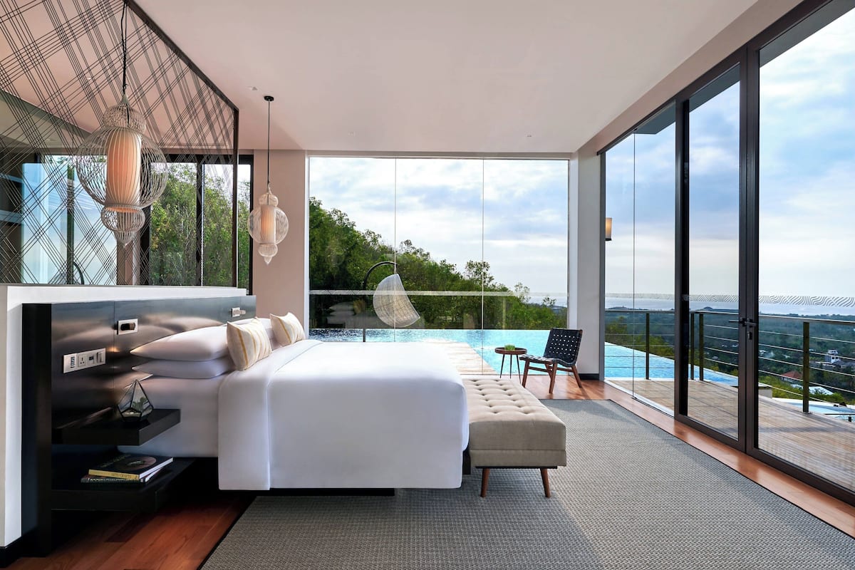 A modern bedroom with a large bed, a nightstand, and a view of a swimming pool and greenery.