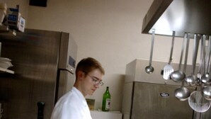 A kitchen with stainless steel appliances, a person in a white coat, and a green bottle on a shelf. - Gasthaus Neupert (Lemberg)