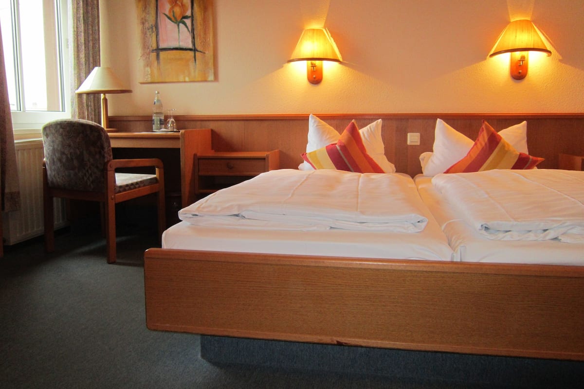A hotel room with a bed, two bedside lamps, a desk, a chair, and a painting on the wall.